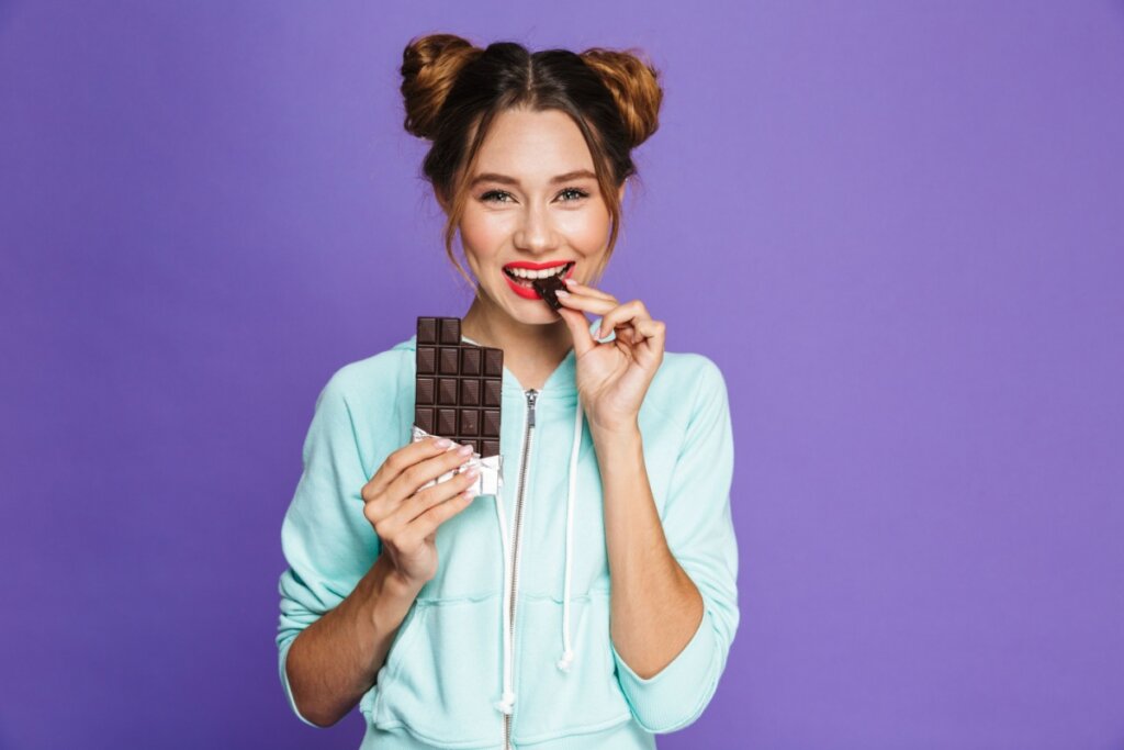 mulher com uma barra de chocolate na mão e com a outra levando um pedaço de chocolate a boca num fundo roxo