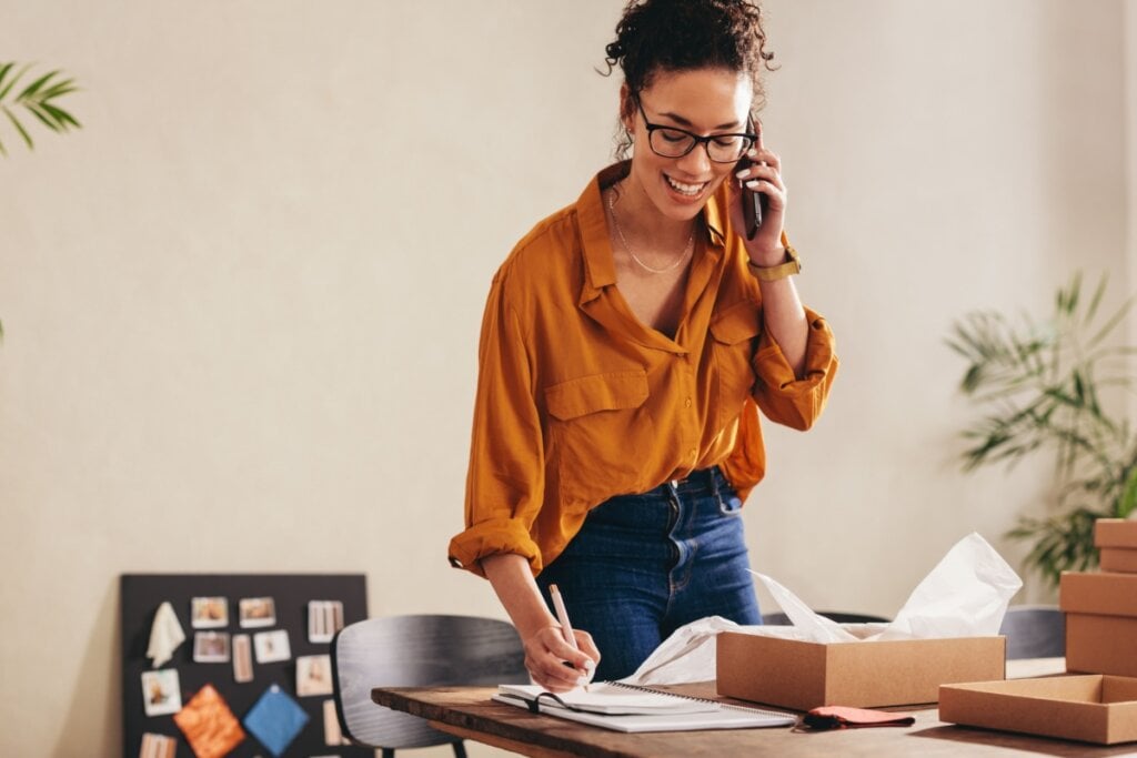 mulher conversando ao telefone e com uma mesa com pedidos para organizar