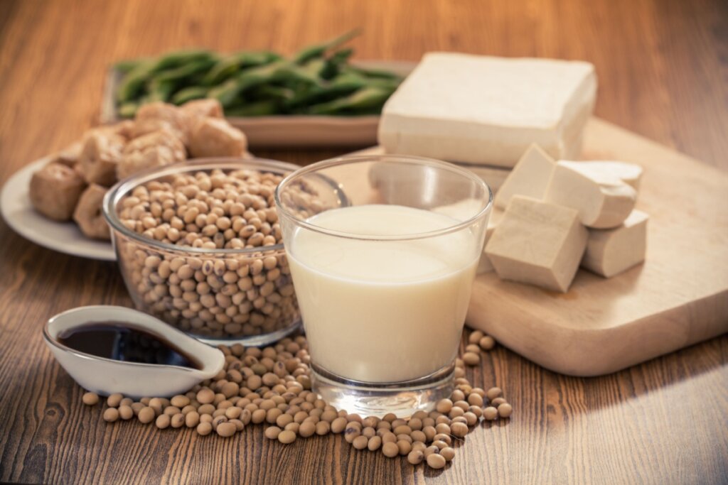 Produtos da soja em cima de uma bancada: copo com leite de soja, pedaços de tofu, uma cumbuca com grãos de soja e uma colher com óleo de soja