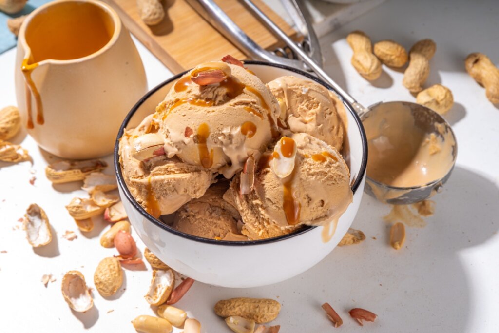 bolas de sorvete de amendoim em uma cumbuca com amendoins espalhados ao redor e talheres em uma bancada
