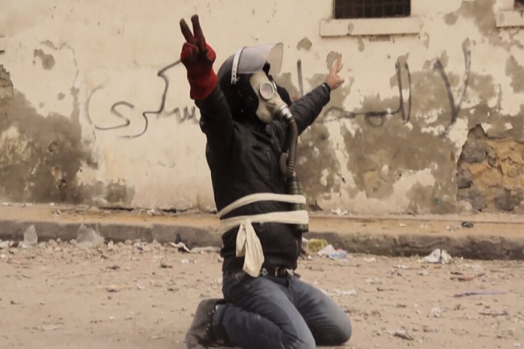 jovem agachado, com roupas de guerra e máscara de gás em manifestação