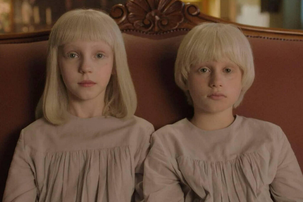 um casal de irmãos gêmeos sentados um ao lado do outro com a mesma roupa e de cabelo loiro 