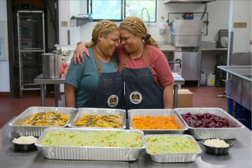 duas irmãs gêmeas se abraçam em frente a uma mesa com diversos formas com comidas diferentes