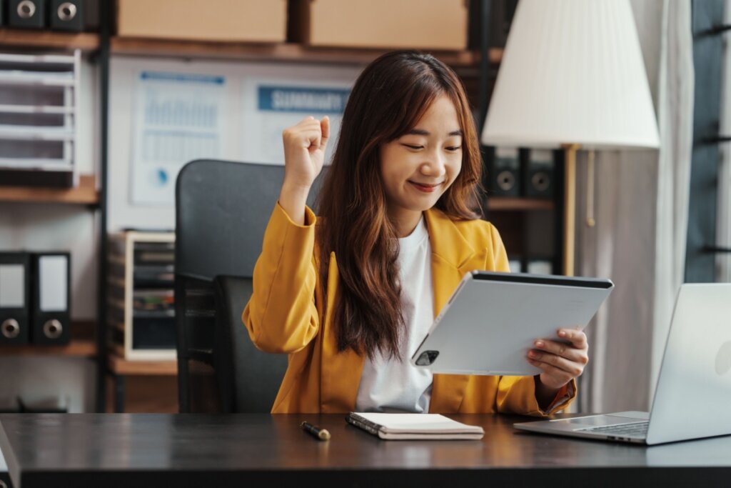 Mulher asiática sentada em uma cadeira em uma sala de escritório com um tablet na mão e comemorando com a outra mão.