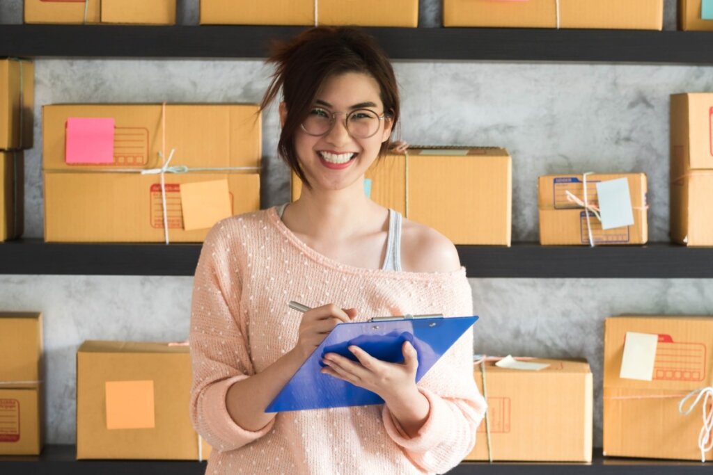 Mulher sorrindo segurando uma prancheta azul em frente a caixas de papelão