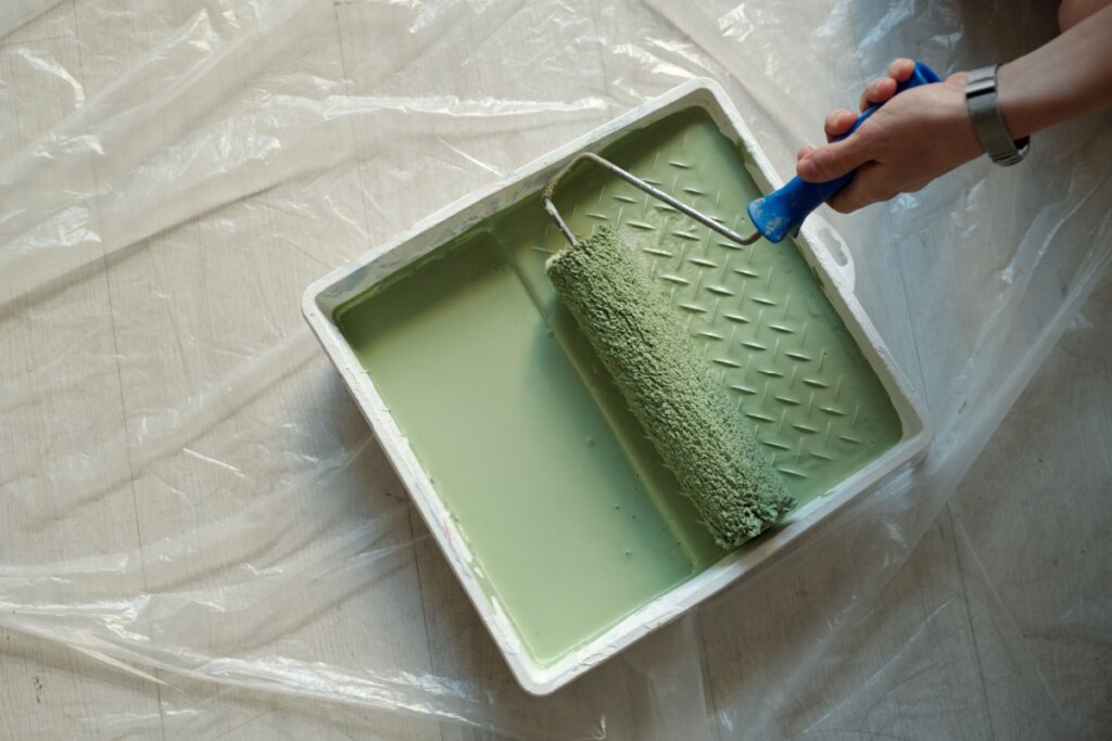 Bandeja de plástico branca com tinta verde no chão coberta com celofane e mão de mulher segurando rolo de pintura