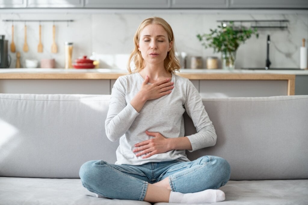 Mulher sentada no sofá com os olhos fechados e as mãos na barriga e no coração