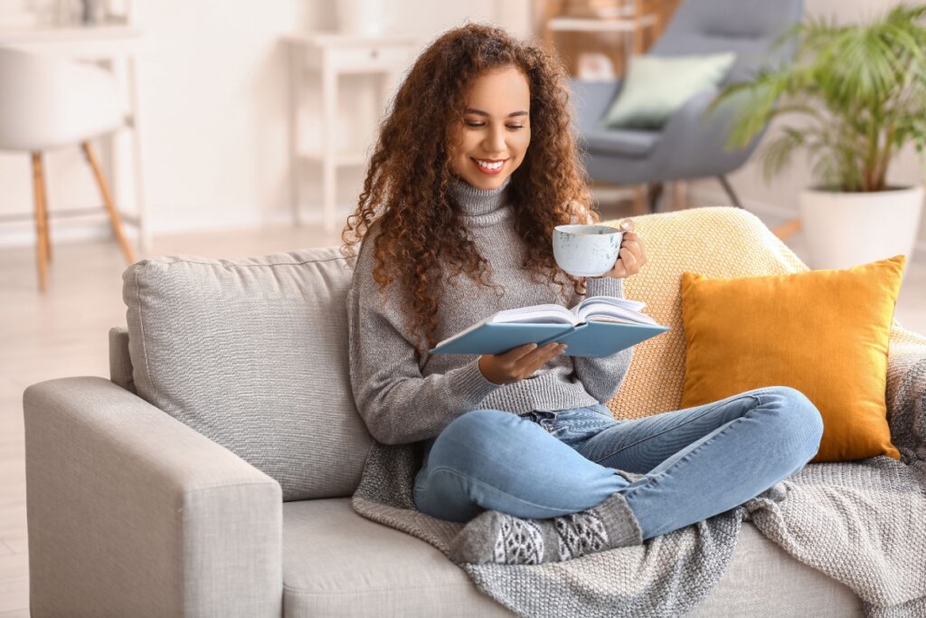 Mulher tomando algo em xícara e lendo livro, sentada em sofá cinza