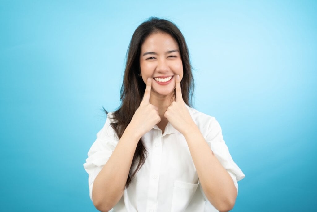 mulher sorrindo indicando o sorriso com as mãos em fundo azul