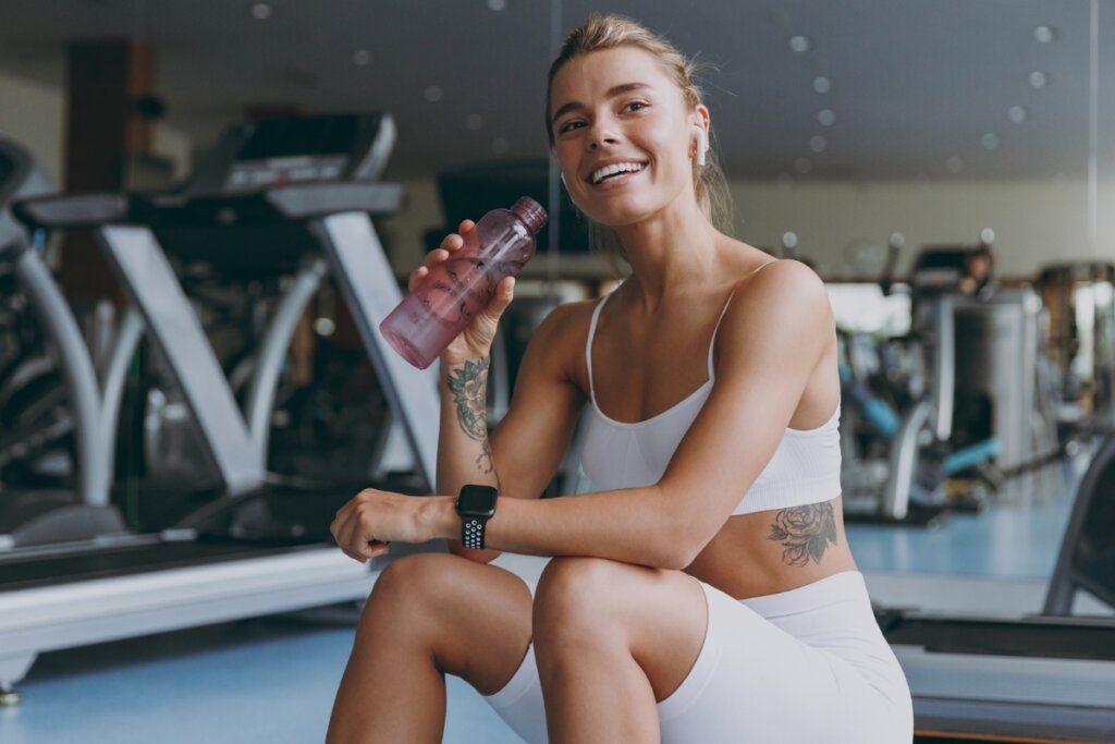 Mulher com roupa de academia branca sentada bebendo água em uma academia