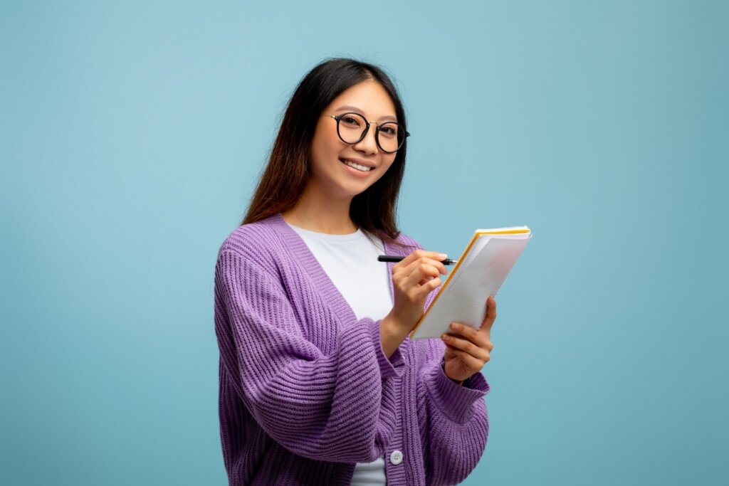 Menina usando óculos preto e um suéter roxo sorrindo e segurando um caderno