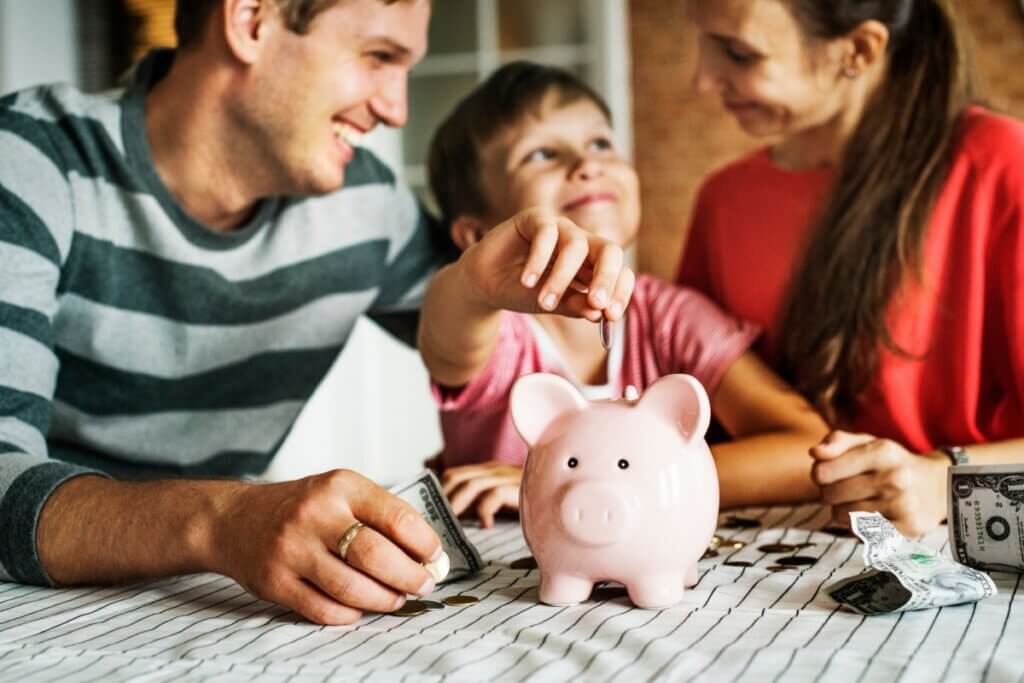 casal com criança, a criança está colocando moedas em um cofre em formato de porco em cima de uma mesa 