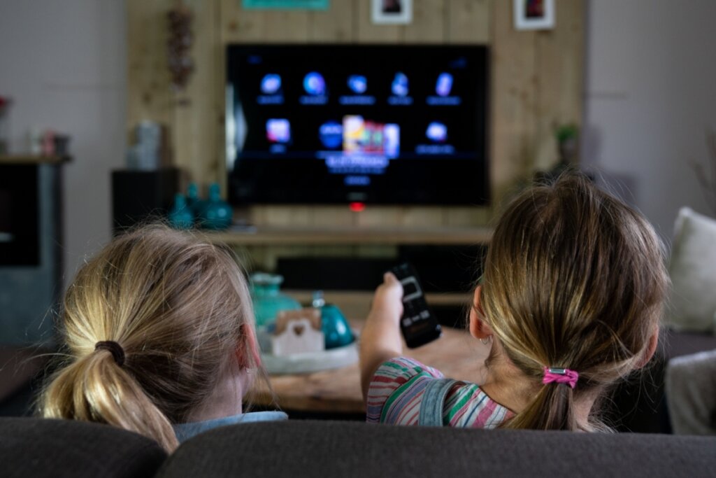 Duas meninas sentadas no sofá da sala com o controle na mão mexendo na televisão