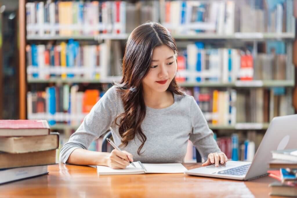 Estudante na biblioteca usando o notebook e escrevendo 