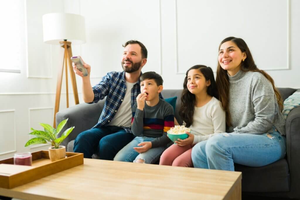 família sentada no sofá com o pai com controle na mão, um casal de crianças comendo pipoca e a mãe assistindo tv