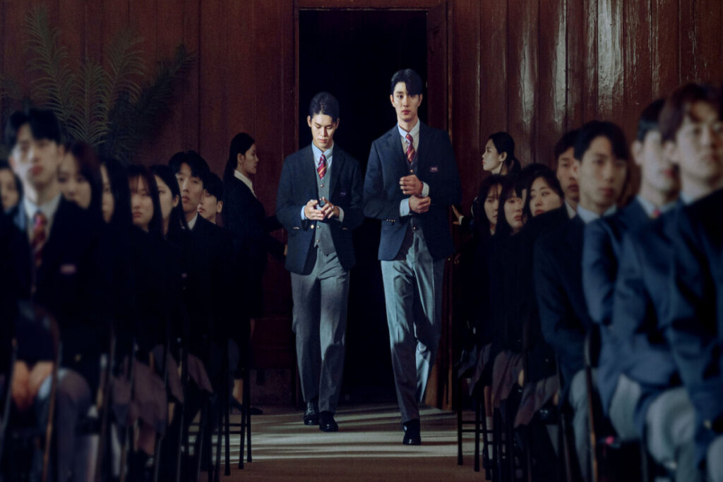 dois alunos no centro da foto andando em um corredor de escola onde há outros alunos sentados