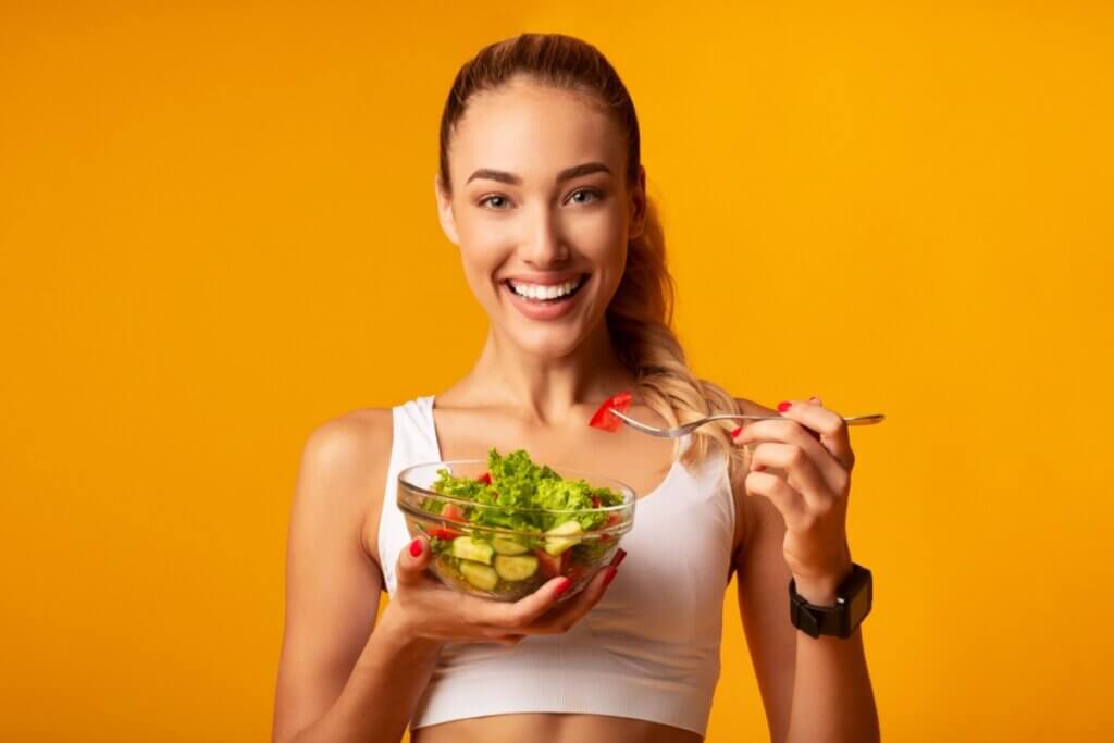 mulher com uma tigela de salada na mão sorrindo e com um tomate em um garfo em fundo laranja