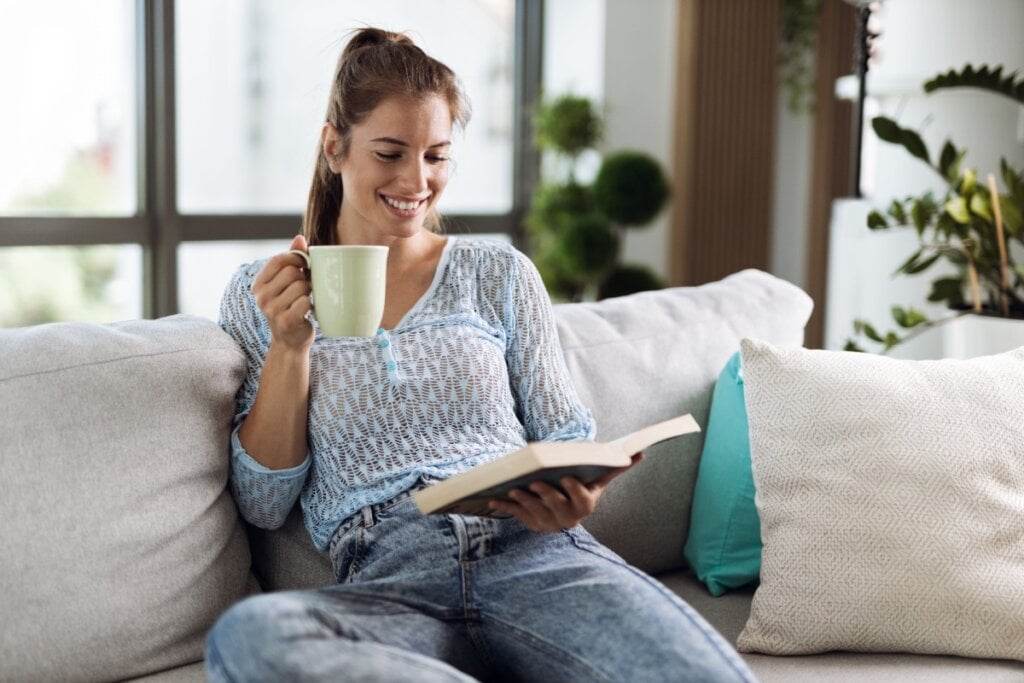 Mulher sentada em um sofá, sorrindo enquanto segura uma caneca e lê um livro, com grandes janelas e plantas ao fundo