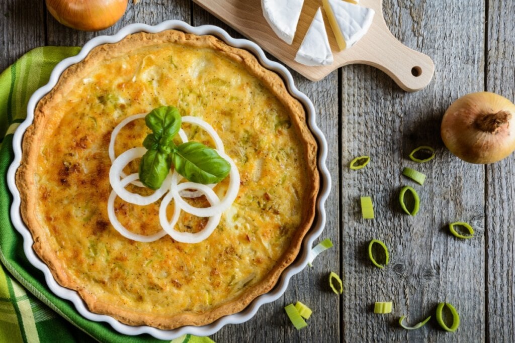 Quiche de alho-poró em refratário branco com rodelas de cebola e folhas de manjericão em cima