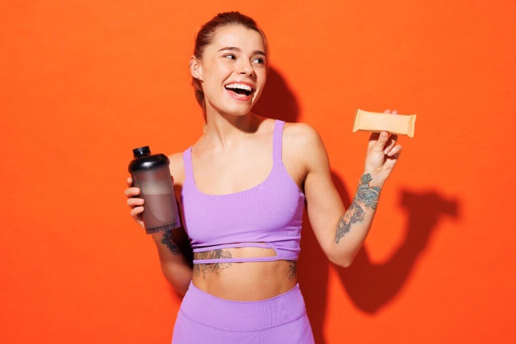 Mulher sorrindo com top roxo segurando garrafa e barrinha de proteína