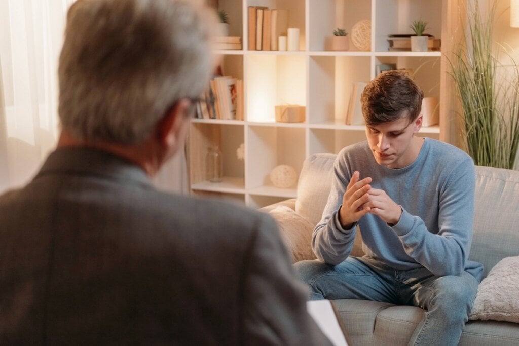 Homem sentado com as mãos unidas e cabeça baixa conversando com um psicólogo