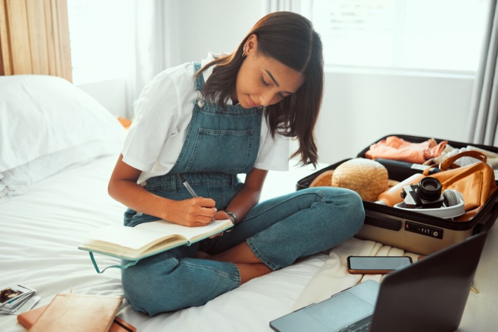 Mulher sentada em uma cama escrevendo em um livro com uma mala com roupas ao lado