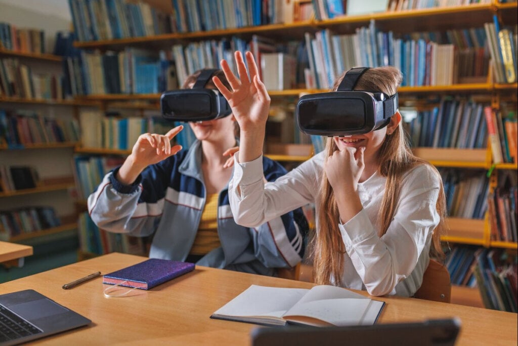 Dois estudantes em uma biblioteca usando óculos de realidade virtual, explorando conteúdos digitais interativos enquanto se divertem e aprendem, com cadernos e notebooks abertos na mesa