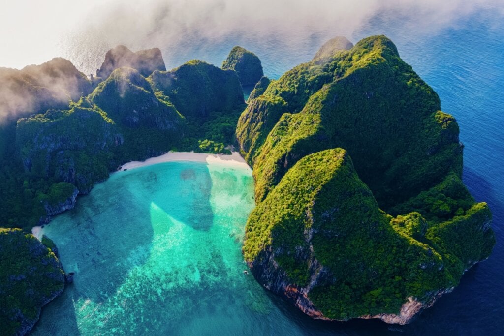 Imagem de cima da ilha Koh Phi Phi na Tailândia, são montanhas cobertas de vegetação e ao meio delas uma faixa de areia com águas cristalinas