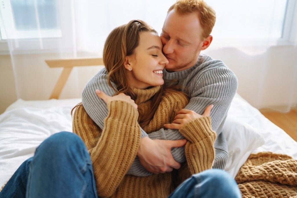 Mulher e homem sentados em uma cama abraçados com o rosto colado