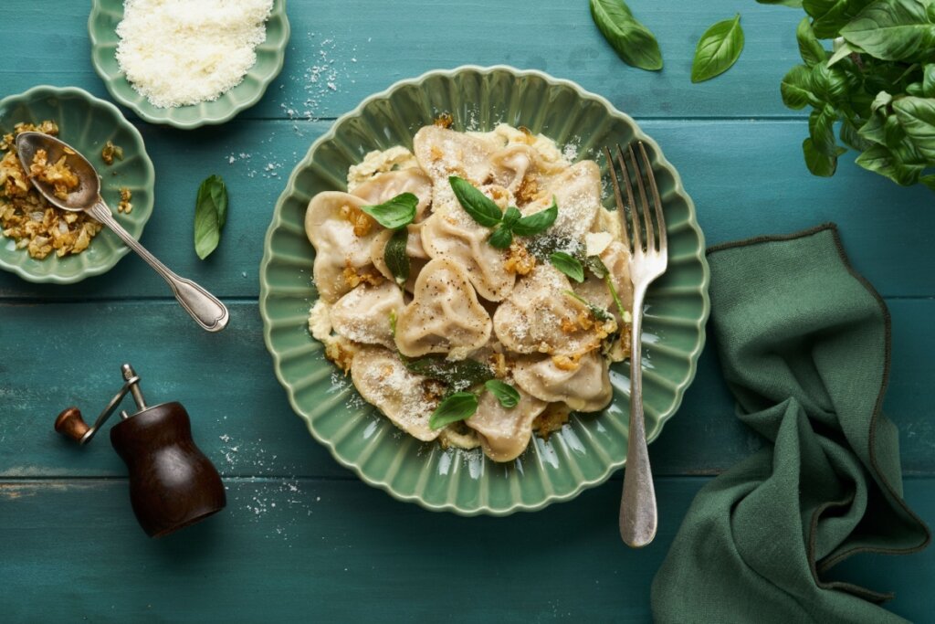 Ravioli caseiro em forma de coração, recheado com carne, cebolas caramelizadas, folhas frescas de manjericão e queijo parmesão ralado, apresentado em um prato verde sobre uma mesa rústica azul