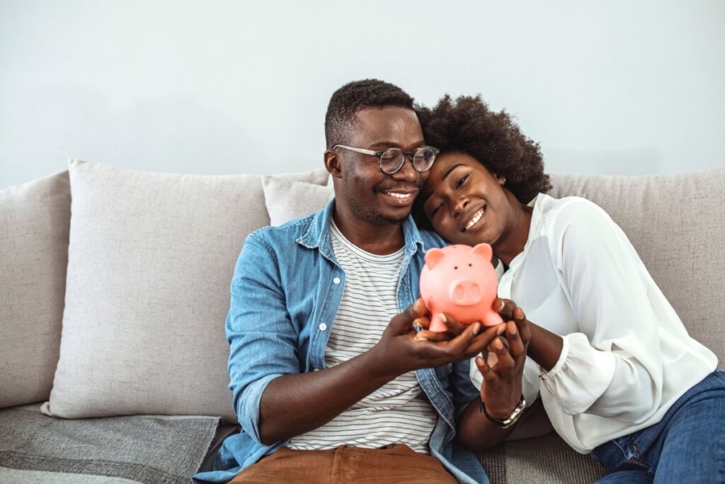 Casal sorrindo, sentados abraçados no sofá segurando um cofrinho em forma de porquinho rosa