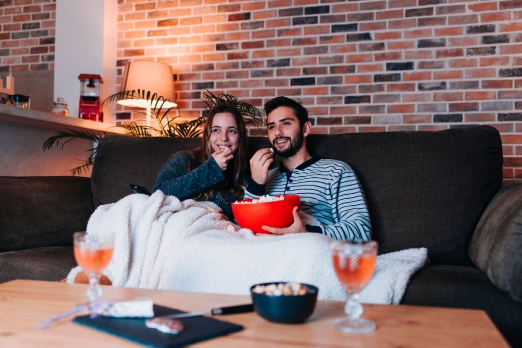 Casal sentado no sofá juntos vendo TV com uma coberta e baldes de pipoca