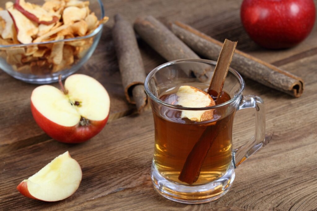 Chá de maçã com canela em xícara transparente, com pedaço da fruta e canela em pau; no fundo, há paus de canela e maçãs cortadas