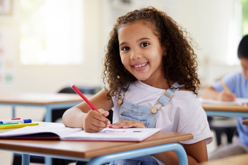 Menina sentada em sala de aula sorrindo para foto e desenhando em caderno