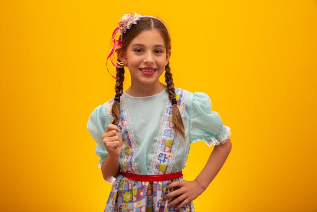 Criança vestida com roupas de festa junina e duas tranças no cabelo, com uma expressão sorridente em um fundo laranja