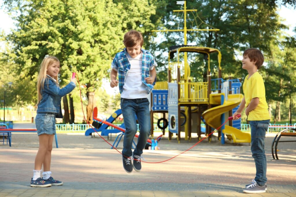 Três crianças brincando no parque, dois segurando uma corda e outro pulando