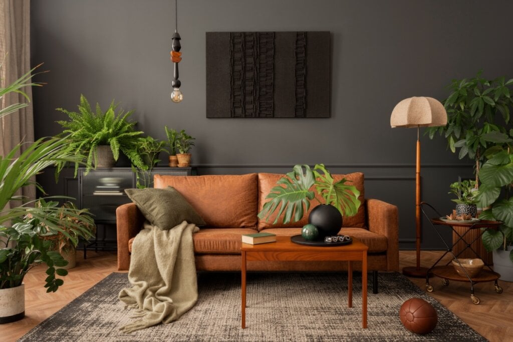 Imagem e uma sala com a parede cinza, um sofá laranja com uma manta e uma almofada verdes. Com diversos vasos de planta 