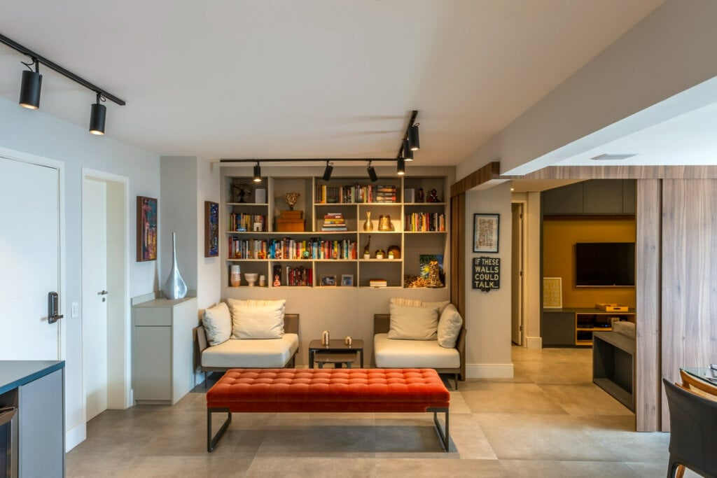 Sala com uma estante ao fundo na parede com nichos e com livros, há duas poltronas brancas e um móvel para sentar com o estofado vermelho