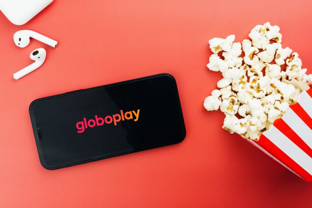 Celular com o logo do Globoplay na tela e ao lado um par de fones de ouvido e um saco com pipoca em um fundo vemelho