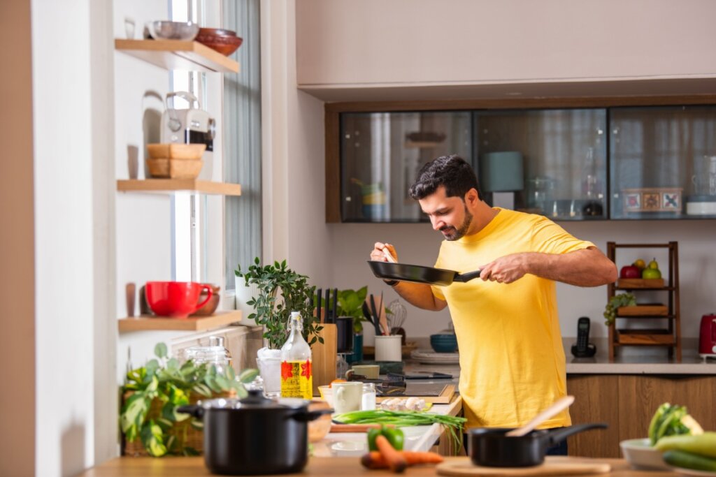 Homem cozinhando, com uma panela na mão sentindo o cheiro do alimento. Ele tem barba e está vestindo uma camiseta amarela