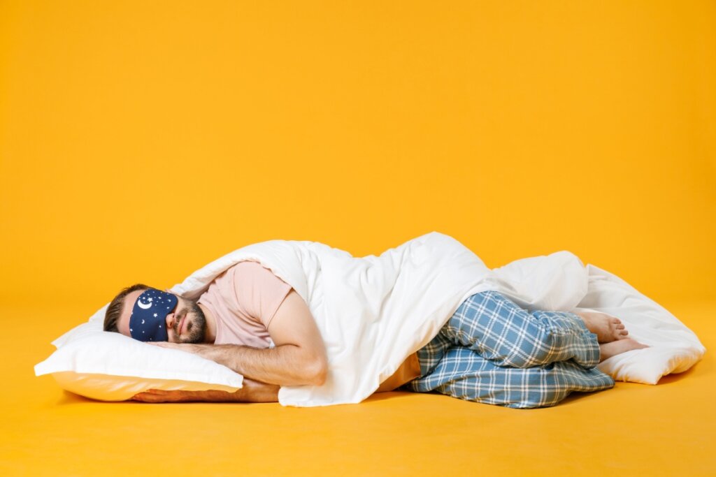 Homem dormindo profundamente em uma cama, vestindo pijamas de flanela azul e uma máscara de dormir estampada com lua e estrelas, contra um fundo amarelo vibrante