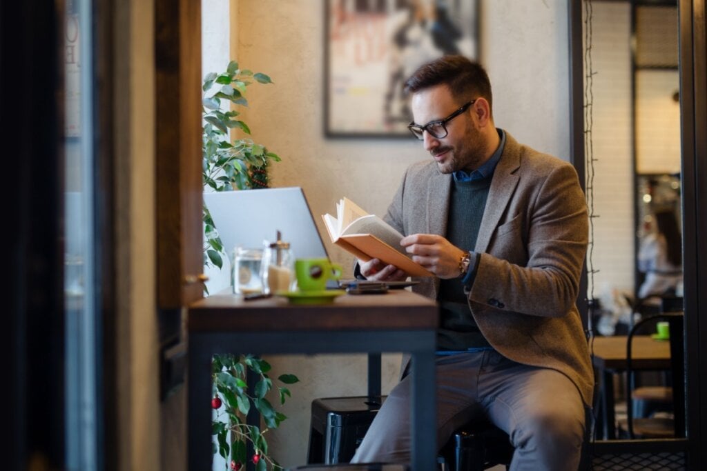 Homem sentado em uma bancada de frente ao computador lendo um livro. Ele está usando óculos preto e tem barba. Está usando um paletó marrom, uma camisa preta e uma calça jeans.