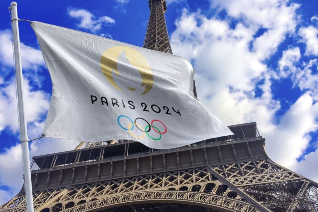 Bandeira dos Jogos Olímpicos de 2024 com o símbolo dos Jogos e também o símbolo do país sede que será Paris, ao fundo está a Torre Eiffel.