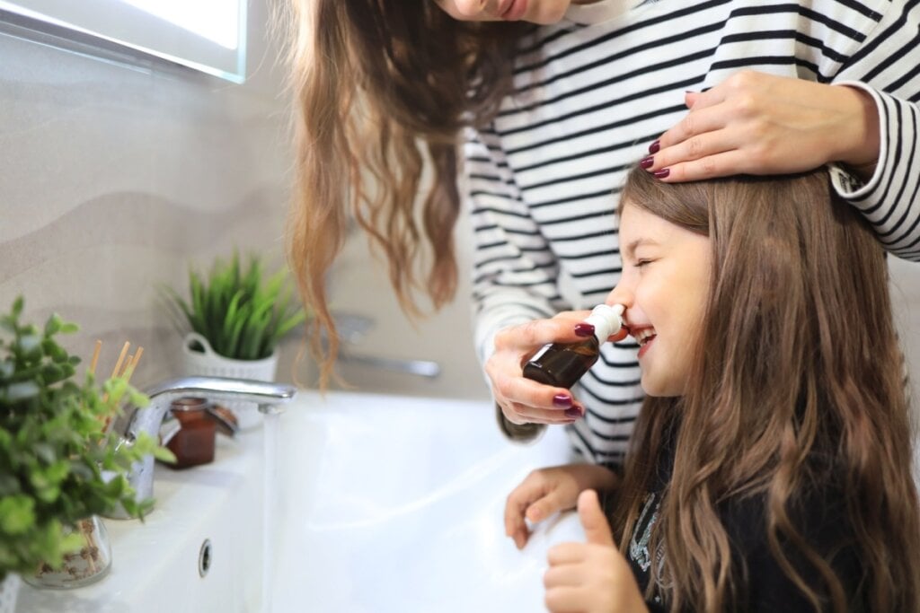 Mãe fazendo uma lavagem nasal na filha na pia do banheiro.