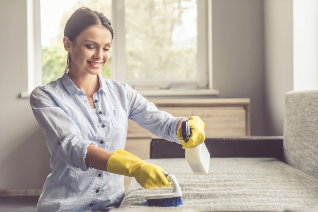 Mulher com uma camisa azul clara de luvas de limpeza, passando uma escova de limpeza branca e cerdas azuis e espirrando produto de limpeza em um sofá cinza-claro