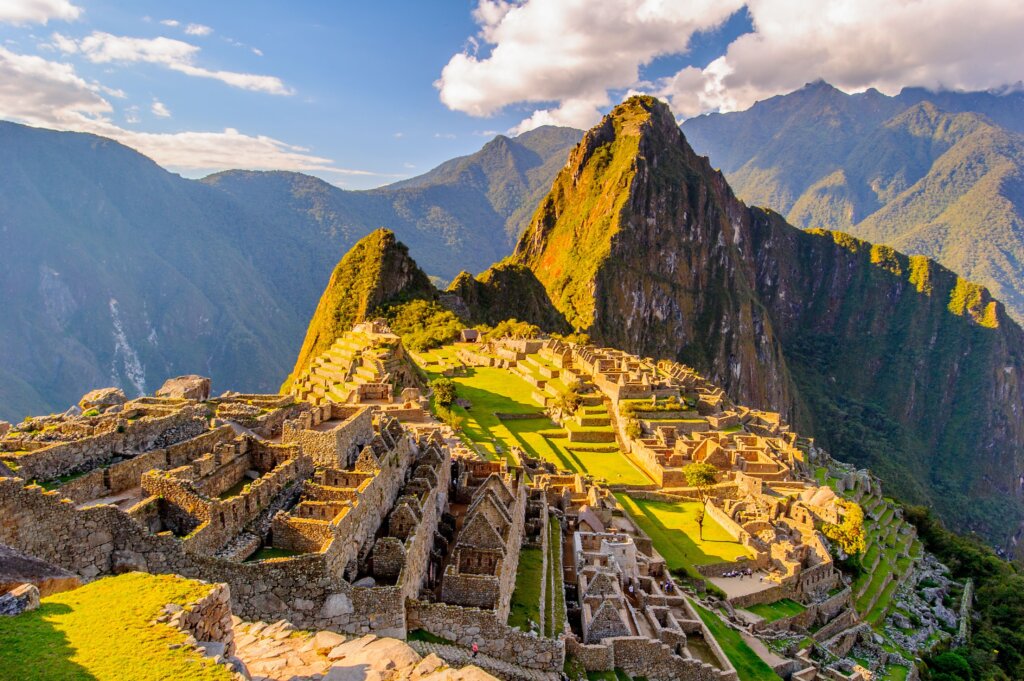 Vista panorâmica de Machu Picchu, a antiga cidade inca situada no alto das montanhas do Peru, com suas ruínas de pedra perfeitamente preservadas e os picos verdes exuberantes ao fundo, iluminados pelo sol da tarde