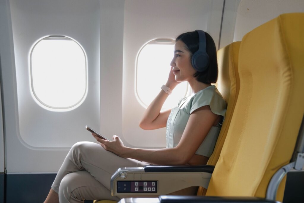 Mulher sentada em poltrona de avião amarela ao lado da janela. Ela usa fone de ouvido e celular