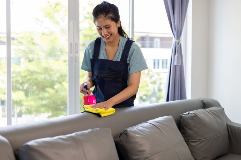 Mulher atrás de sofá limpando ele com produto rosa e pano amarelo