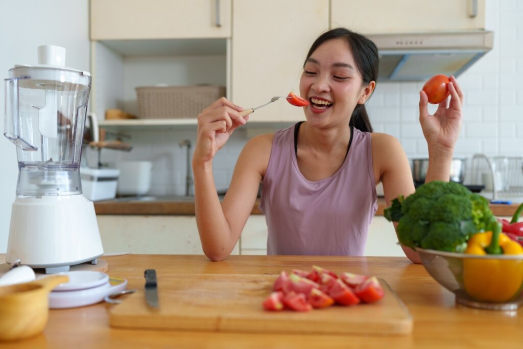 Mulher comendo tomates em uma bancada de madeira. Na bancada está uma bandeja de corte com uma faca e pedaços de tomar, ao lado há uma cesta com outros alimentos. 