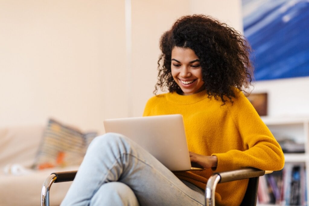 Mulher fazendo pesquisa no computador, ela está usando um suéter amarelo e uma calça jeans, o cabelo está solto e ela está sorrindo